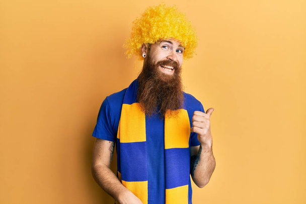Rotschopf Mann mit langem Bart Fußball Hooligan Anfeuerungsspiel mit lustiger Perücke lächelnd mit glücklichem Gesicht und zeigt zur Seite mit erhobenem Daumen.  - Foto, Bild