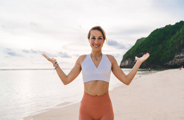 Mulher positiva em pé à beira-mar com os braços para cima enquanto pratica asana durante o treino de fitness na costa arenosa e olhando para a câmera - Foto, Imagem