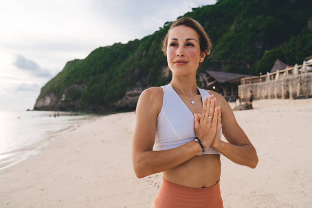 Slim female in trendy activewear showing namaste gesture and thoughtfully looking away against tropical shore with sandy beach and calm ocean in daylight - Fotografie, Obrázek