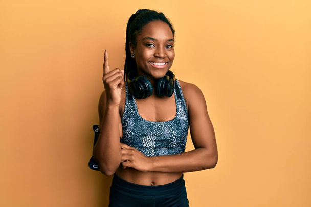 African american woman with braided hair wearing sportswear and arm band with a big smile on face, pointing with hand and finger to the side looking at the camera.  - Fotoğraf, Görsel
