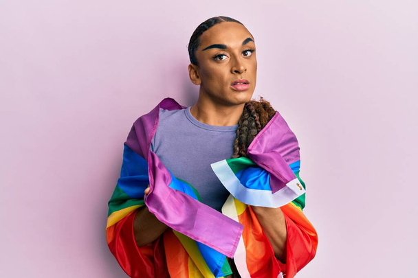 Hispanic man wearing make up and long hair wrapped in rainbow lgbtq flag clueless and confused expression. doubt concept.  - Photo, Image