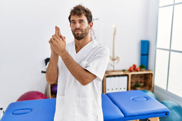 Young handsome physiotherapist man working at pain recovery clinic holding symbolic gun with hand gesture, playing killing shooting weapons, angry face  - Fotoğraf, Görsel