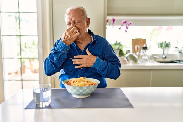 Homme âgé aux cheveux gris mangeant des spaghettis aux pâtes à la maison sentant quelque chose de puant et dégoûtant, odeur intolérable, retenant son souffle avec les doigts sur le nez. mauvaise odeur  - Photo, image