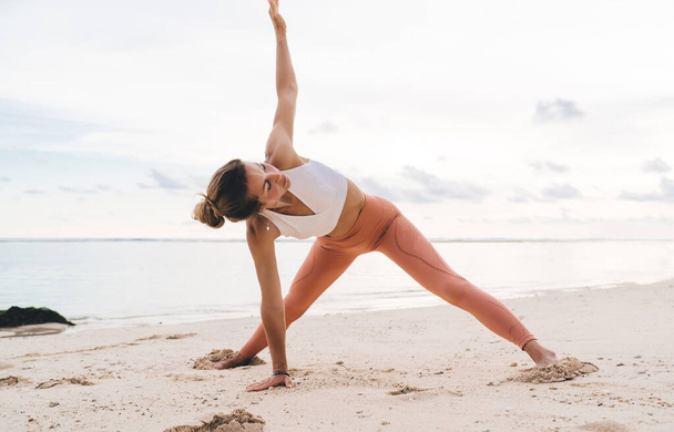 Flexible slender female with hair bun in crop top and leggings doing Trikonasana while practicing yoga on quiet seashore in Bali - Φωτογραφία, εικόνα