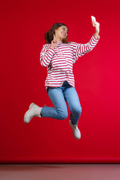 Emotional young beautiful girl in sweater and jeans taking selfie isolated on red studio background. Concept of emotions, facial expression - Foto, immagini