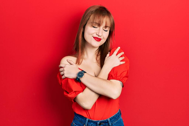 Redhead young woman wearing casual red t shirt hugging oneself happy and positive, smiling confident. self love and self care  - 写真・画像