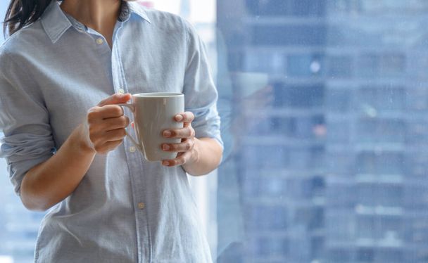 Kadın eli, arka planda şehir manzarası bulanık olan beyaz bir fincan kahve ya da çay tutuyor. Boşluğu kopyala - Fotoğraf, Görsel