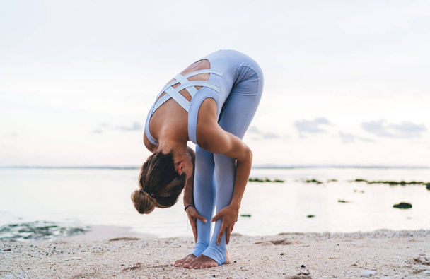 Flexible female athlete in active wear practicing yoga for healthy body and mind, sportive fit girl doing asanas feeling zen and harmony during morning training workout at coastline near ocean - Foto, Imagen