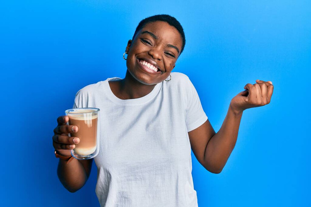 Młoda Afroamerykanka trzymająca kawę krzyczy dumna, świętując zwycięstwo i sukces bardzo podekscytowana podniesionym ramieniem  - Zdjęcie, obraz