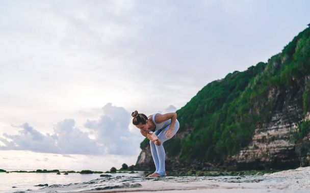 From below of full length of young female doing yoga exercise on rocky coast near sea in evening summer time - Фото, изображение