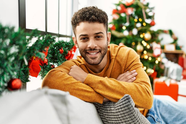 Young arab man smiling happy sitting on the sofa by christmas tree at home. - Foto, imagen