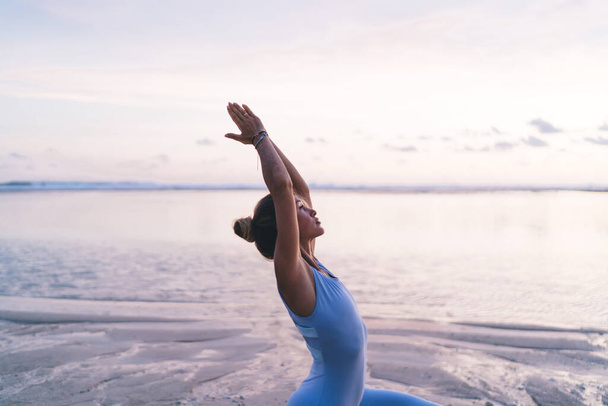 Side view of concentrated ethnic female athlete in sport bodysuit raising folded hands while training yoga pose alone against vibrant sunrise sky and ocean water - Φωτογραφία, εικόνα