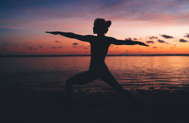 Silhouette of slim female athlete with perfect body shape training flexibility of muscles during evening twilight, active girl doing asanas at yoga workout enjoying physical recreation at seashore - Photo, Image
