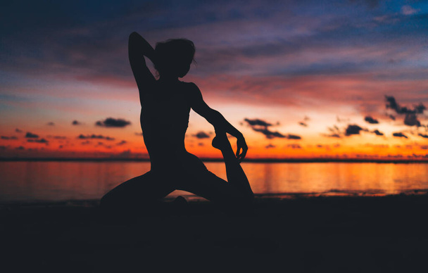 Silhouette of anonymous woman sitting in Crooked Monkey pose and throwing hand behind head while doing yoga asana against calm sea during colorful sundown - Photo, Image