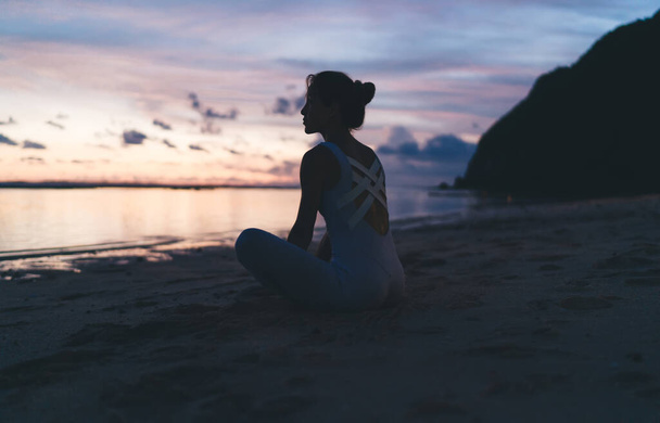 Back view of young woman in sportswear sitting in lotus pose on sandy beach against cloudy sky over calm sea in evening - Fotoğraf, Görsel