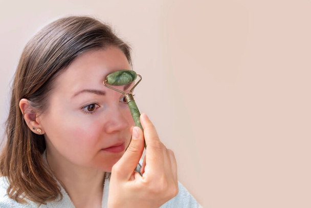 Jovem mulher caucasiana fazendo massagem facial com rolo de jade verde no fundo bege - Foto, Imagem