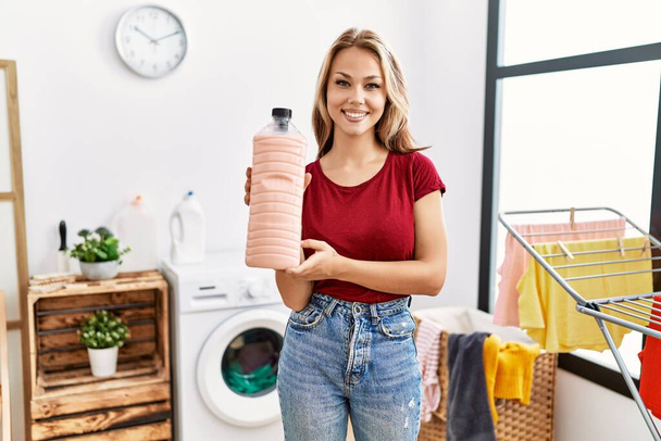 Young caucasian woman holding detergent bottle at laundry room looking positive and happy standing and smiling with a confident smile showing teeth  - Foto, Imagen