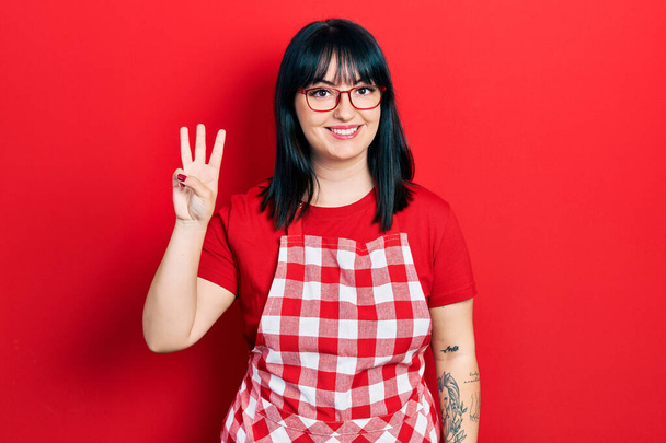 Mujer hispana joven usando delantal de cocinero y gafas mostrando y apuntando hacia arriba con los dedos número tres mientras sonríe confiado y feliz.  - Foto, imagen