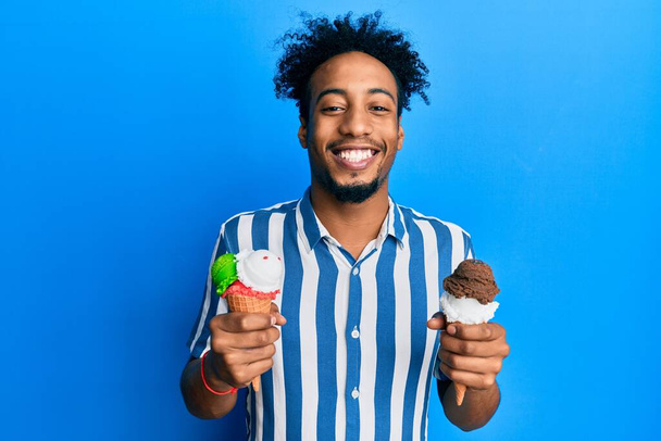 Jovem afro-americano com barba segurando dois cones de sorvete sorrindo com um sorriso feliz e legal no rosto. mostrando dentes.  - Foto, Imagem