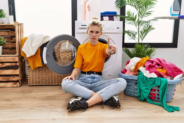 Junge blonde Frau beim Wäschewaschen sitzt an der Waschmaschine und zeigt mit erhobenem Finger und wütendem Gesichtsausdruck, zeigt keine Geste  - Foto, Bild