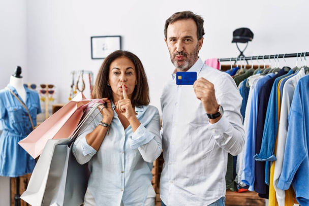 Latijns-Amerikaans echtpaar van middelbare leeftijd met boodschappentassen en kredietkaart die vragen om stil te zijn met de vinger op de lippen. stilte en geheim concept.  - Foto, afbeelding