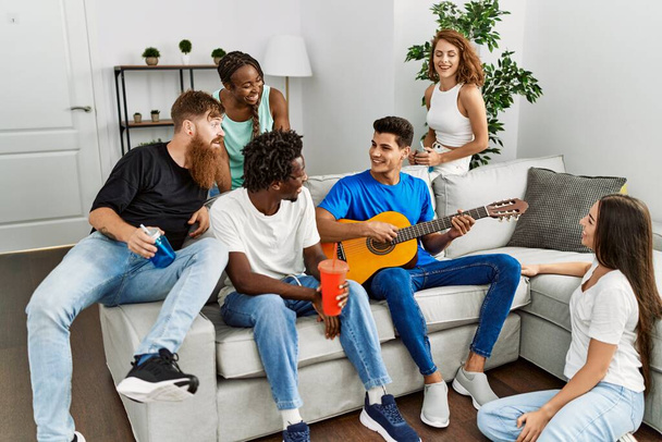 Groep jonge vrienden die vrolijk glimlachen en thuis klassieke gitaar spelen. - Foto, afbeelding