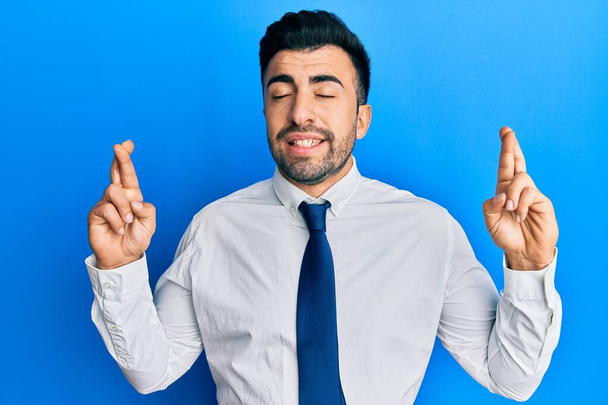 Young hispanic man wearing business clothes gesturing finger crossed smiling with hope and eyes closed. luck and superstitious concept.  - Фото, изображение
