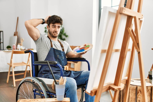 Young hispanic man sitting on wheelchair painting at art studio confuse and wondering about question. uncertain with doubt, thinking with hand on head. pensive concept.  - Foto, imagen