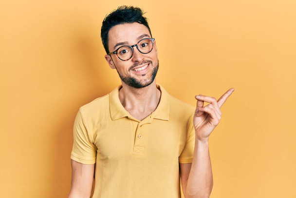 Fiatal spanyol férfi alkalmi ruhát és szemüveget visel, nagy mosollyal az arcán, kezével és ujjával a kamera felé mutat..  - Fotó, kép