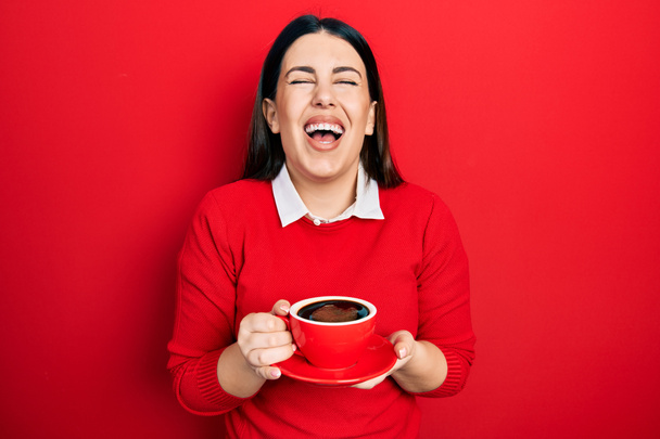 Mujer hispana joven bebiendo una taza de café sonriendo y riendo en voz alta porque divertida broma loca.  - Foto, imagen