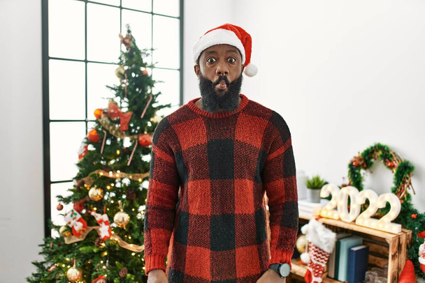 Αφροαμερικάνος που φοράει καπέλο Σάντα Κλάους και στέκεται δίπλα στο χριστουγεννιάτικο δέντρο κάνοντας γκριμάτσες με χείλη, τρελή και κωμική χειρονομία. αστεία έκφραση.  - Φωτογραφία, εικόνα