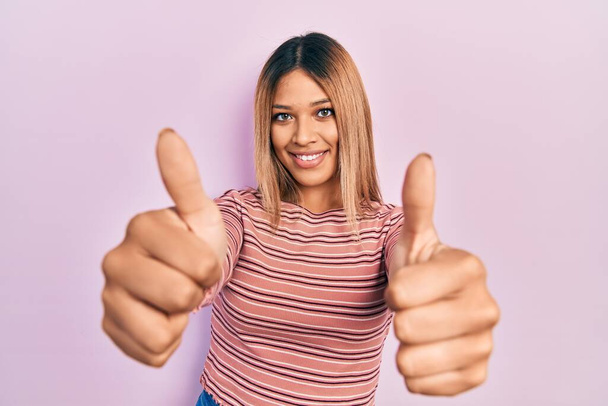 Beautiful hispanic woman wearing casual striped sweater approving doing positive gesture with hand, thumbs up smiling and happy for success. winner gesture.  - Foto, Bild