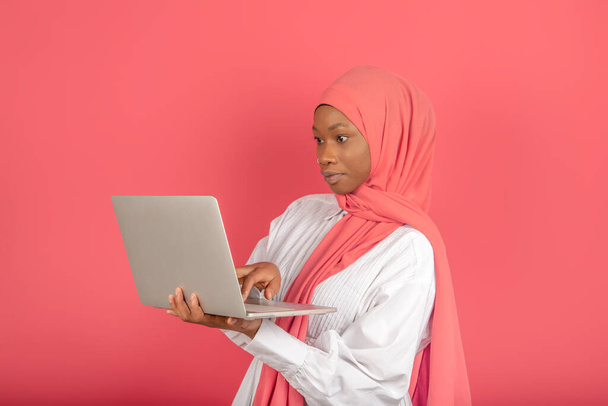 Muslim college student working concentrated on laptop standing on pink background - Foto, Imagen