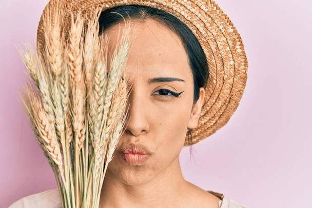 Young hispanic girl wearing summer hat holding spike wheat over eye looking at the camera blowing a kiss being lovely and sexy. love expression.  - Foto, immagini