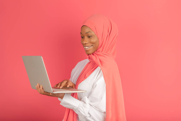 Portret studentki pracującej na laptopie. Piękna młoda, czarna afrykańska muzułmanka z różowym hidżabem w różowym studio. Stała ze szczęśliwym uśmiechem na twarzy.. - Zdjęcie, obraz