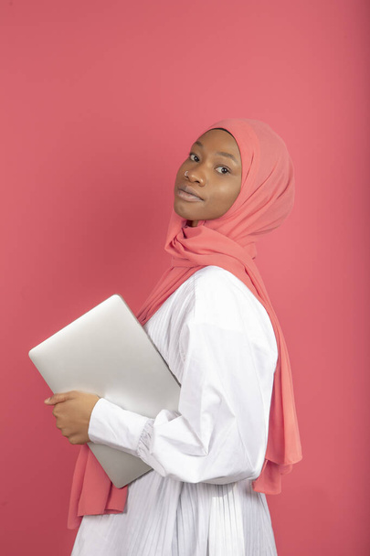 Uma jovem afro-americana de perfil vestindo lenço rosa e segurando um laptop isolado no fundo rosa - Foto, Imagem