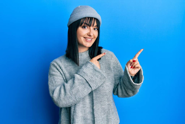 Young hispanic woman wearing wool sweater and winter hat smiling and looking at the camera pointing with two hands and fingers to the side.  - Photo, Image