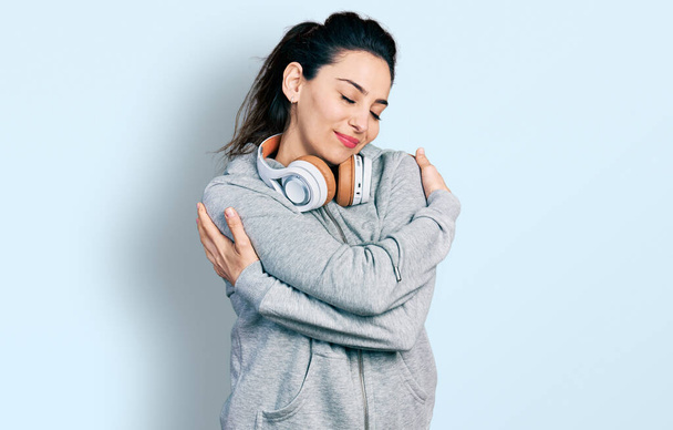 Mujer hispana joven vistiendo ropa de gimnasia y usando auriculares abrazándose feliz y positivo, sonriendo confiado. amor propio y cuidado personal  - Foto, Imagen