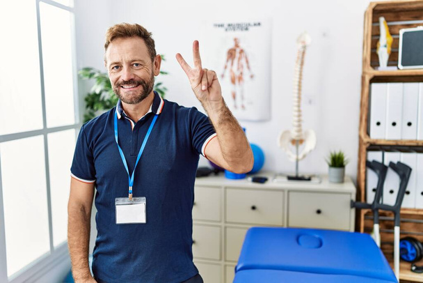 Középkorú fizioterapeuta, aki a fájdalomcsillapító klinikán dolgozik, és a második ujjal mutogat, miközben magabiztosan és boldogan mosolyog..  - Fotó, kép