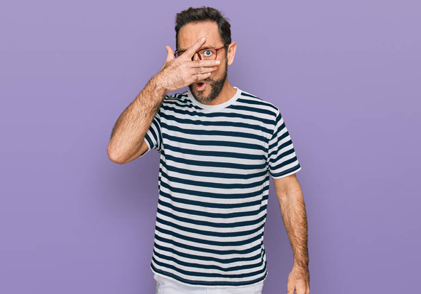 Middle age man wearing casual clothes and glasses peeking in shock covering face and eyes with hand, looking through fingers with embarrassed expression.  - Foto, Imagem