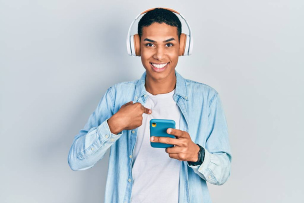Giovane ragazzo afroamericano utilizzando smartphone con le cuffie che puntano il dito verso un sé sorridente felice e orgoglioso  - Foto, immagini
