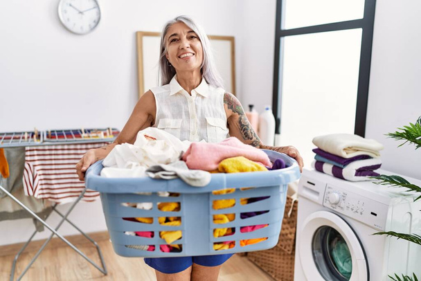 Middle age grey-haired woman holding laundry basket smiling with a happy and cool smile on face. showing teeth.  - 写真・画像