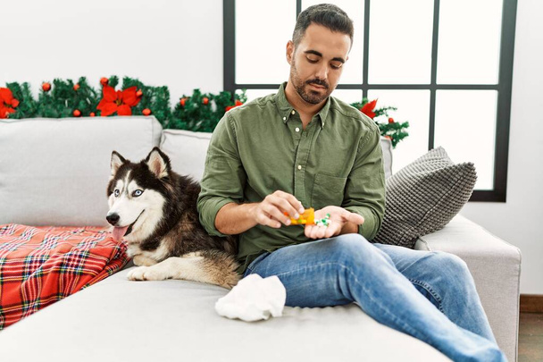 Joven hombre hispano tomando píldoras sentado en el sofá con el perro por la decoración de Navidad en casa - Foto, Imagen