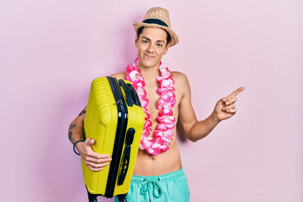 Young hispanic man wearing summer hat and holding cabin bag smiling happy pointing with hand and finger to the side  - Foto, imagen