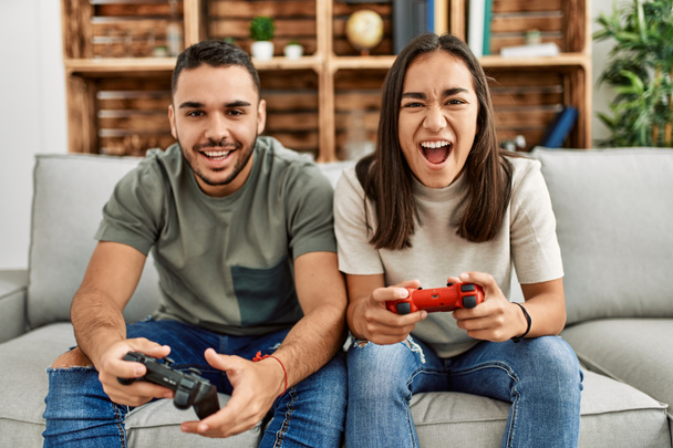 Νεαρό ζευγάρι Λατίνων χαμογελά ευτυχισμένος παίζοντας βιντεοπαιχνίδι στο σπίτι. - Φωτογραφία, εικόνα