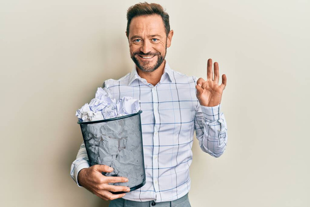 Hombre de mediana edad sosteniendo un cubo de papel lleno de papeles arrugados haciendo buen signo con los dedos, sonriendo gesto amistoso excelente símbolo  - Foto, imagen