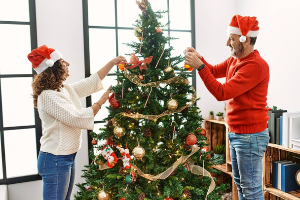 Middelbare leeftijd hispanic paar glimlachen gelukkig versieren kerstboom thuis. - Foto, afbeelding