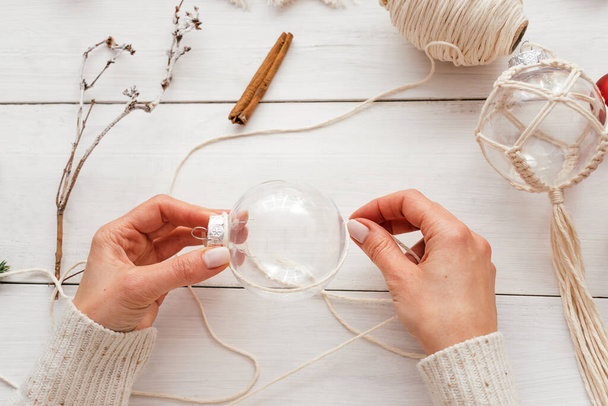 A woman decorates a transparent Christmas ball with macrame-style weaving,handmade decor in eco-style,hands close-up,top view.Christmas, New Year and eco-friendly concept. - Foto, Bild