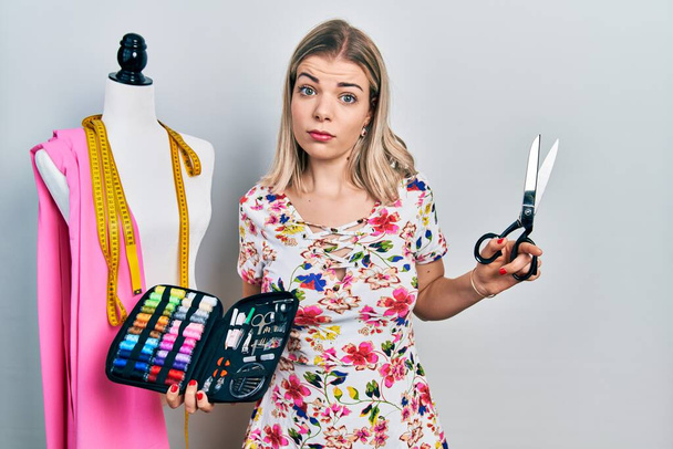 Beautiful caucasian woman dressmaker designer holding scissors and sewing kit clueless and confused expression. doubt concept.  - Foto, Bild