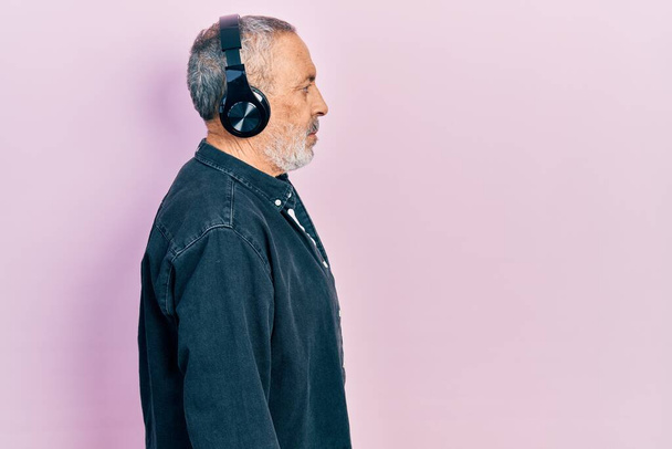 Handsome senior man with beard listening to music using headphones looking to side, relax profile pose with natural face with confident smile.  - 写真・画像